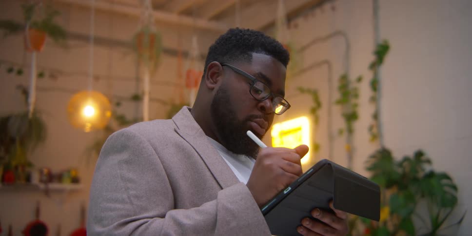 A person writing on a notepad and stylus in a cozy room with hanging plants and warm lighting