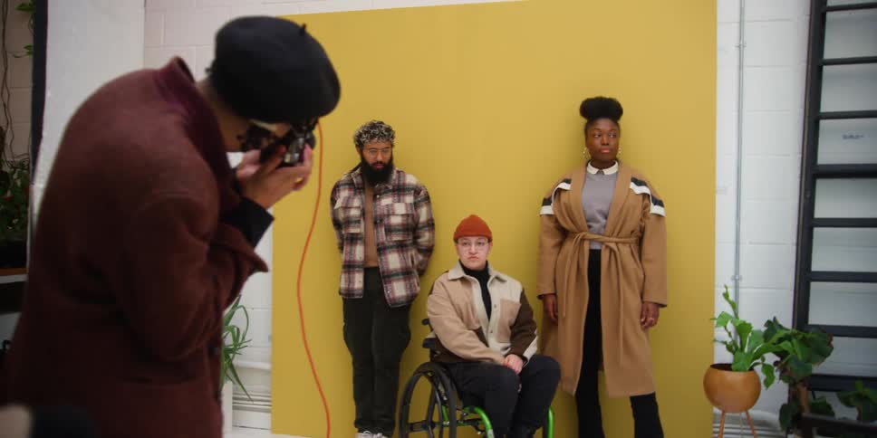 A man taking pictures of three people standing close to a yellow wall with one sitting on a wheelchair