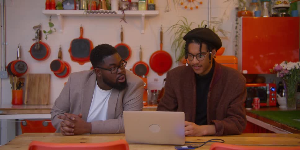 Two black men sitting and talking inside the kitchen with one operating the computer
