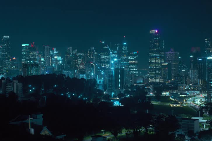 Construction cranes glow against city in background as they build a skyscraper at night