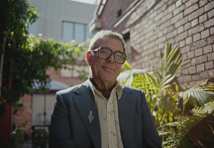 A man wearing glasses smirks while standing in an alley
