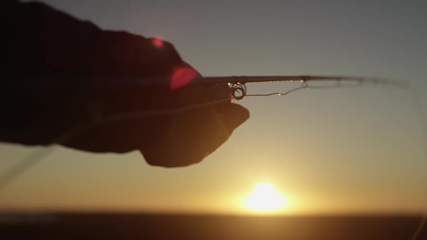 A human hand holding a fishing rod and the sun is strong in the horizon