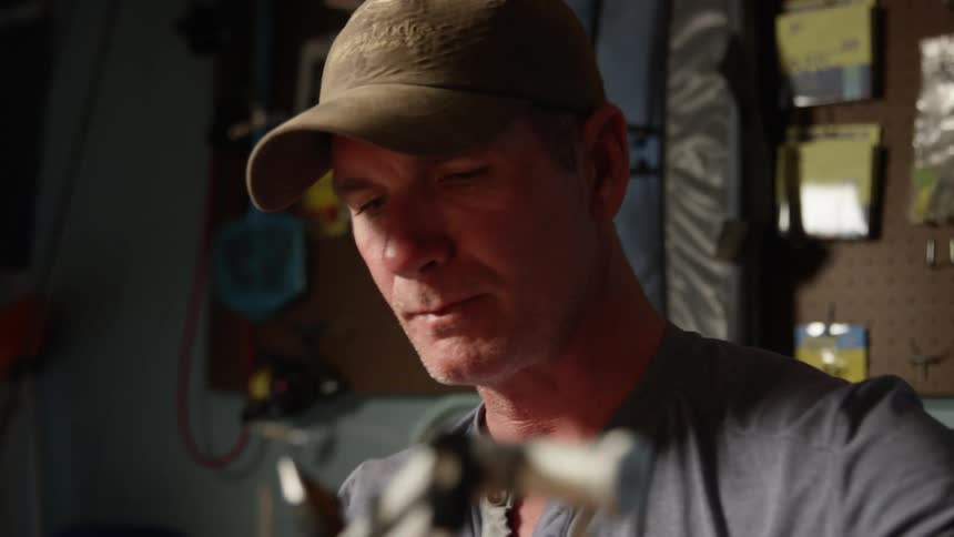 A man in a cap drinks a liquid from a glass and then begins to pass a line through a thin or metallic object attached to a support