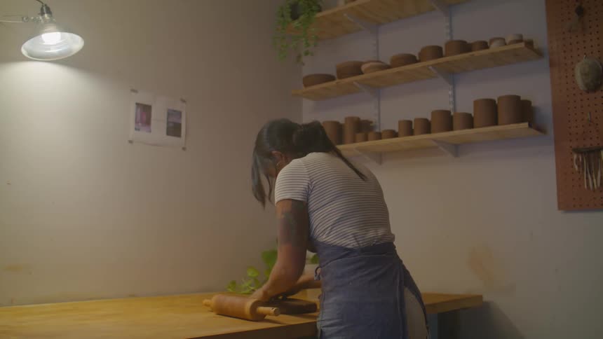Woman indoors standing up next to a wooden table kneading dough with her hands and with a rolling pin