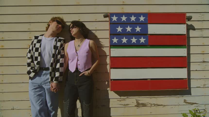 Man and woman side by side holding hands leaning against the wall of a wooden house
