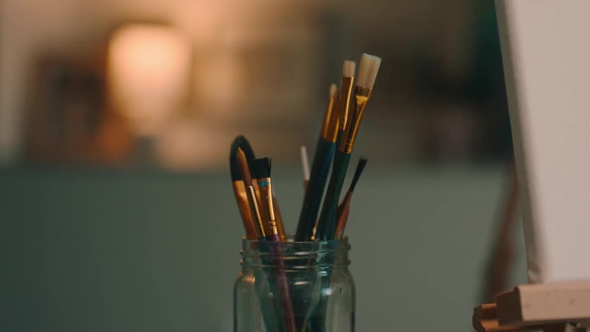 Glass jar next to canvas and easel holds several paintbrushes