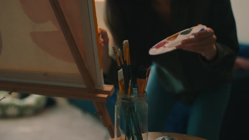 Closeup of a jar with brushes with a woman painting a canvas next to it