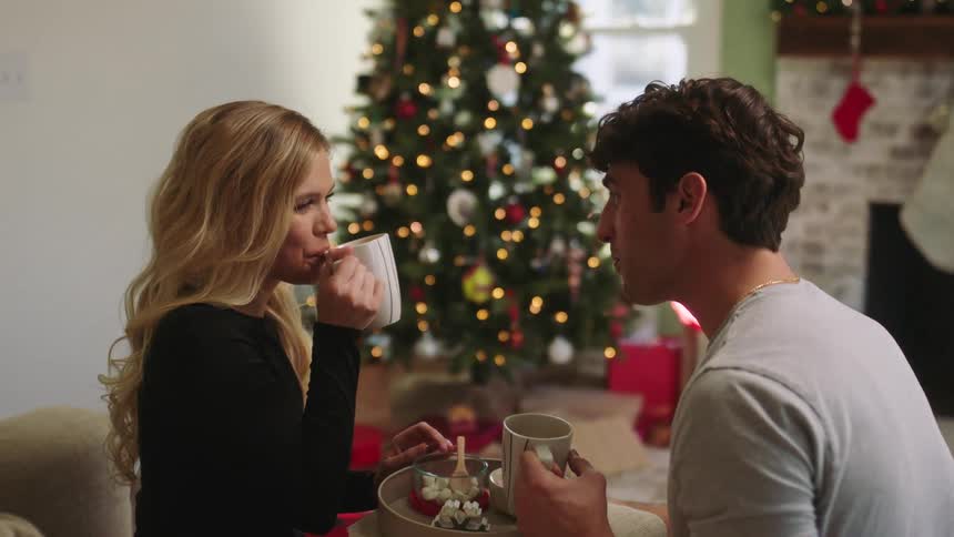 A young couple drink from mugs in a living room at christmastime