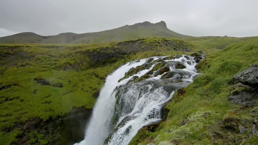 Flowing river creating a small waterfall amidst mosscovered hills on a cloudy day
