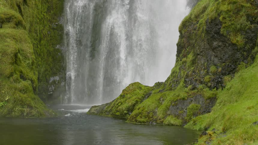 Waterfall flows down moss covered cliff into river pool creating heavy mist