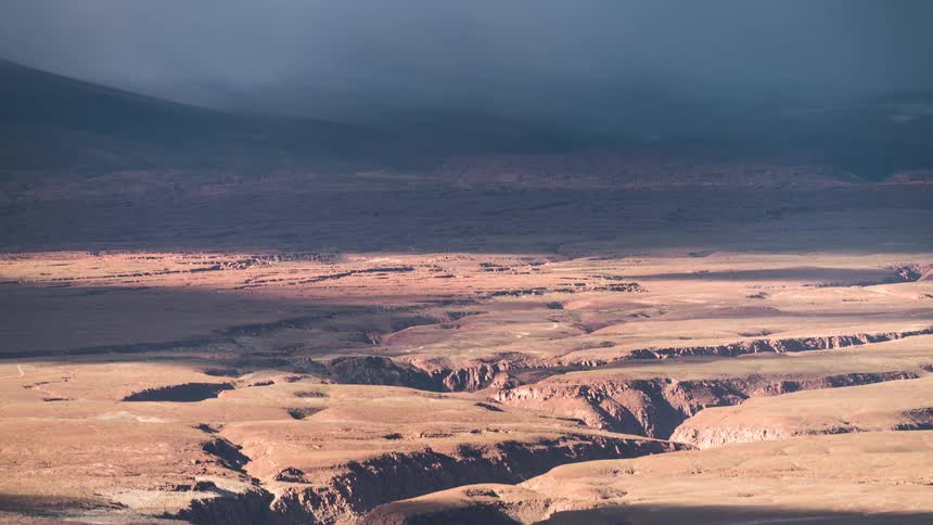 Clouds passing over arid desert crossed by ravines cast shadows over both desert and nearby mountainous terrain