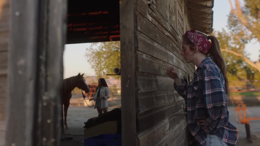 Woman wearing flannel shirt leans against wooden barn and wipes face