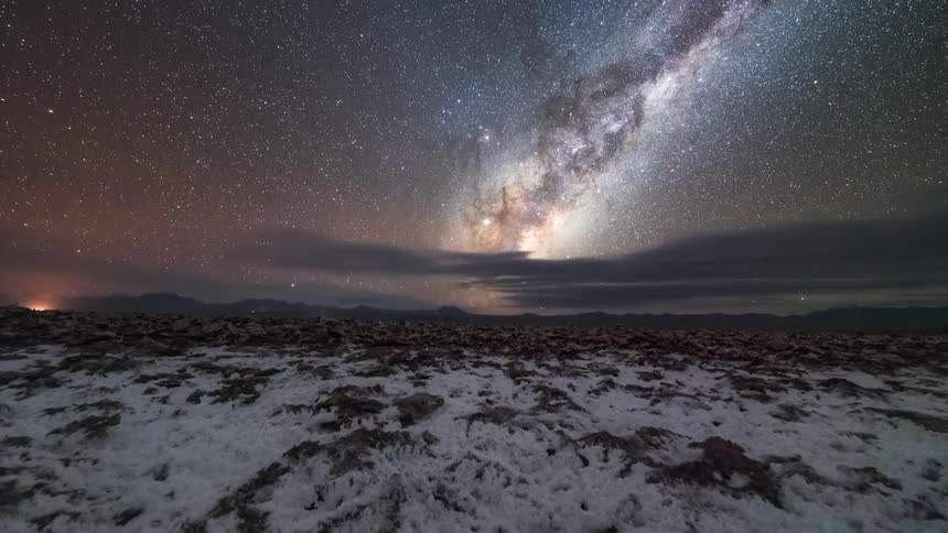 The sun rises in the sky behind the milky way over a rocky snow dusted landscape