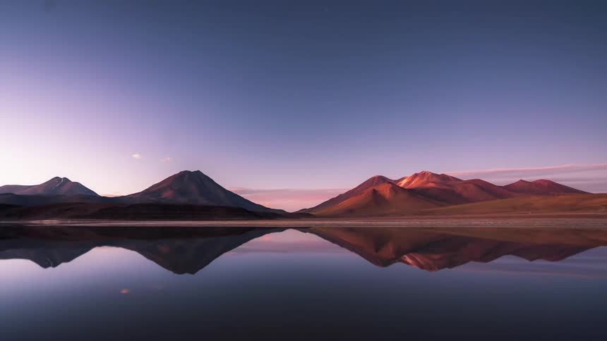 Natural landscape of a reflective lake with mountains around and the sky dawning from a nebula and darkening