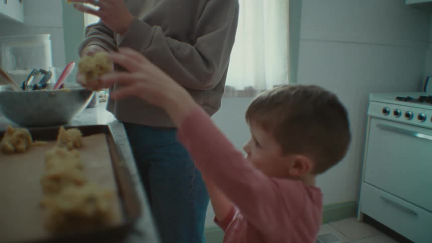 A mother hands her young son a clump of cookie dough to put on a baking pan