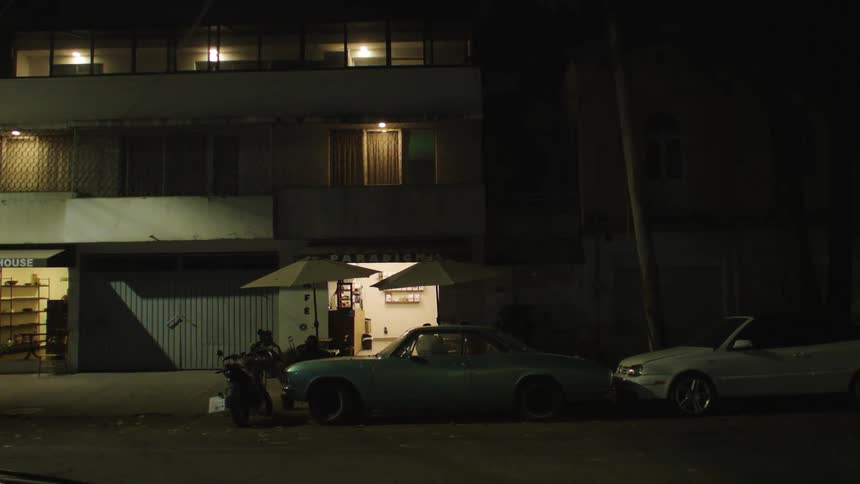 Two cars parked in front of apartment buildings with sun umbrellas and coffee tables still outside at night
