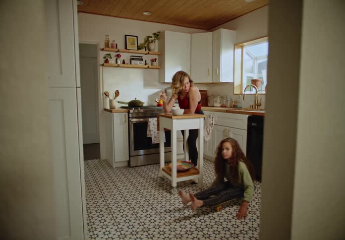 Mother leans against kitchen island eating while daughter rolls across floor on skateboard