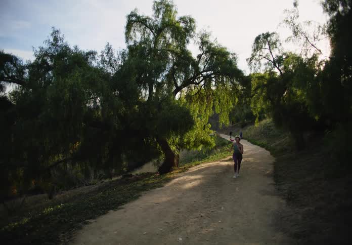 Female runner runs along dirt path through woods while people walk dogs in the background