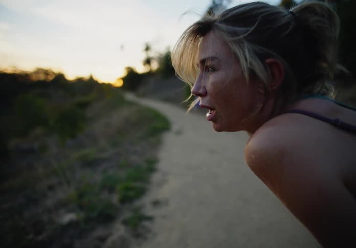 A tired woman stretches on a dirt path during sunset with their hands behind their head