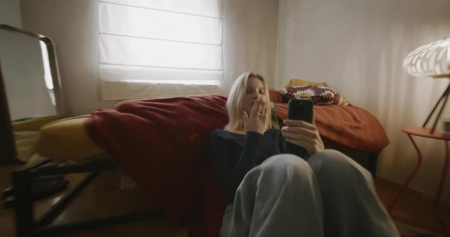 Blonde woman wearing a blue shirt and jeans sits by bed making a video call on smartphone