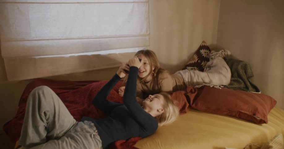 Two girls in a bed laugh while looking at a phone
