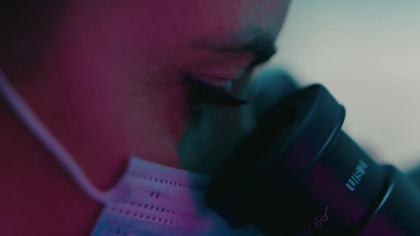 A womans face with a lab mask leans in to look into a microscope