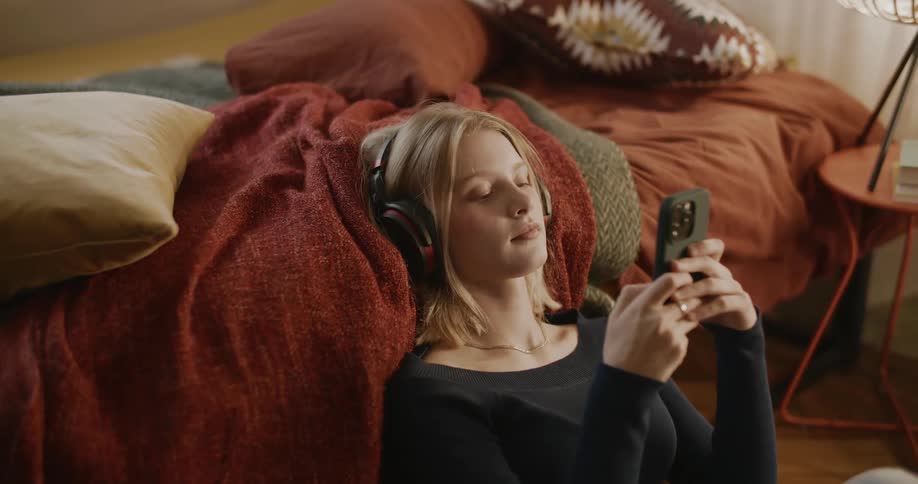 A teenage girl sitting on the floor of her room her back against the side of her bed with her headphones on while she scrolls on her smart phone