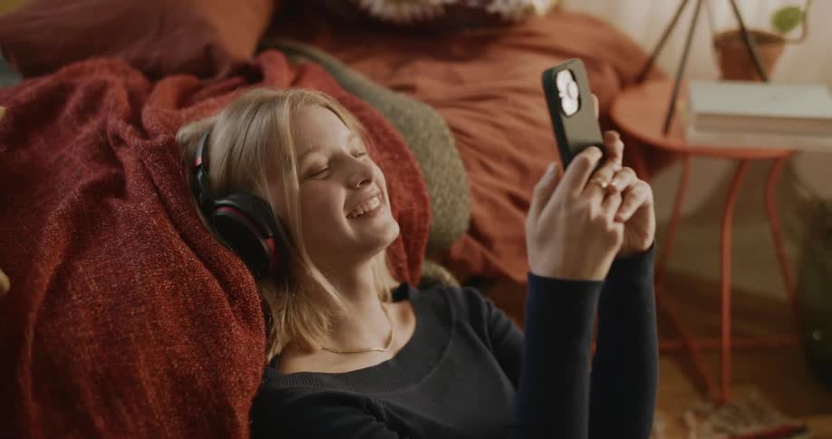 Portrait of a young woman sitting next to the bed smiling using her cell phone and using headphones