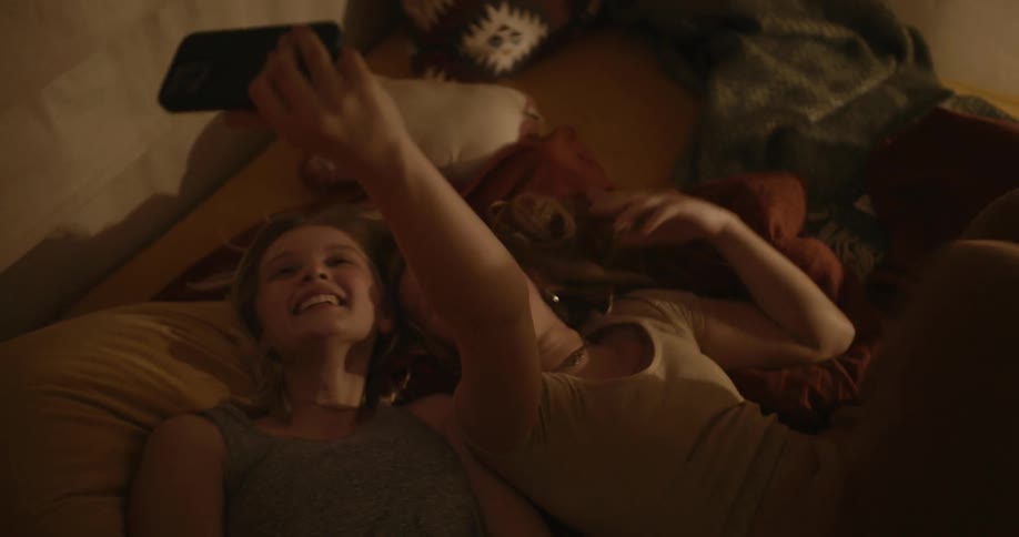 Two young girls are lying on a bed holding a smartphone while gesturing for a selfie