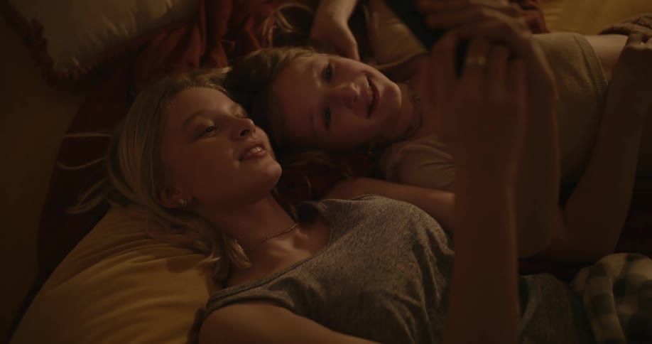 Two teenage girls on a bed looking up at a smart phone while they laugh