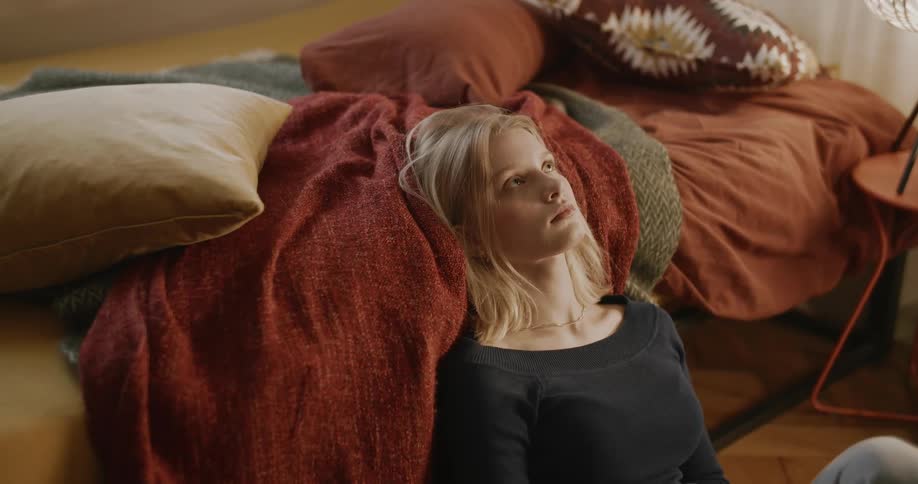 A woman sits at the side on a bed with various pillows and blankets