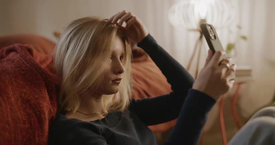 Young blonde woman fixing her hair and smiling at her cellphone