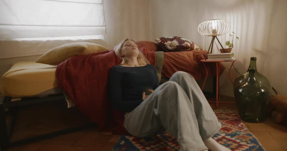 A teenage girl holding a phone leans back against a bed and closes her eyes