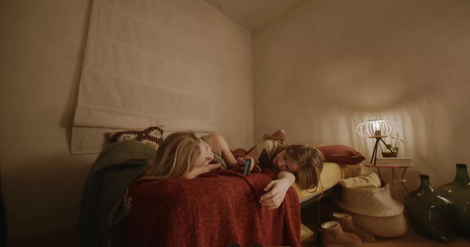 Two female teenagers lay on the bed talking while holding mobile phones