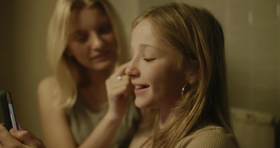 Woman uses smartphone to view herself while friend helps her apply makeup