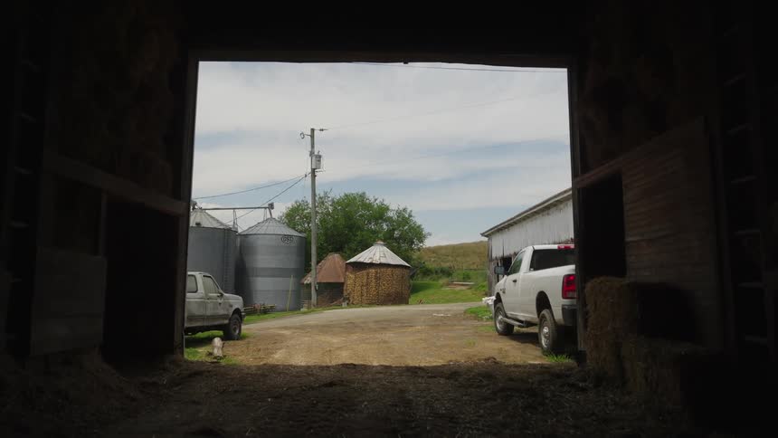 A view from inside a barn to a rural scene with two vehicles and a silo