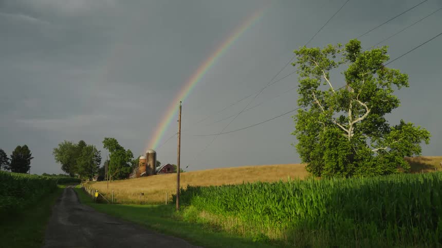 A rainbow brightens the cloudy sky above a countryside scene with a dirt road and a barn