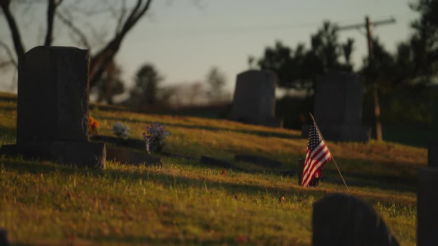 A serene cemetery at sunset with gravestones casting long shadows and an american flag marking one grave