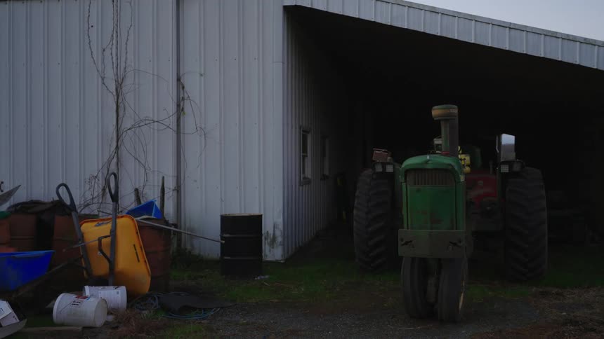 A green tractor is parked outside a large white barn under an overcast sky
