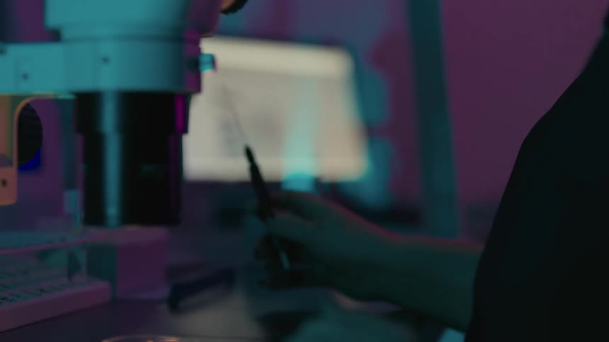 Woman looking through a microscope and simultaneously injecting into a small analysis vial with a needle