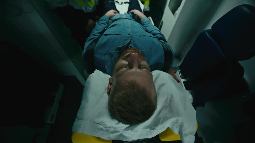 A man is lying unconscious on a stretcher and two people place him inside a vehicle and open his shirt