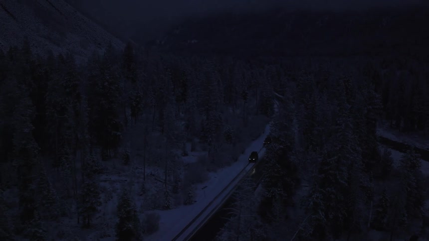 A pair of vehicles travel along a remote road through a snowy forest