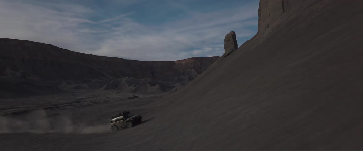 Vehicle drives across desert and base of arid mountain slope creating dust cloud