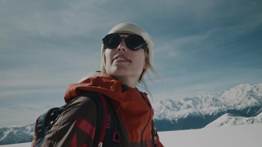 A blonde woman wearing sunglasses and warm clothes while walking in a snowy environment with mountains in the background