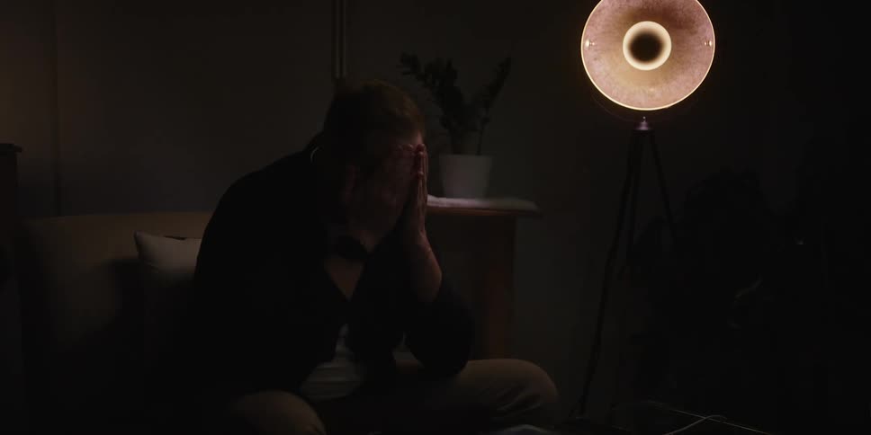 A sad man sitting alone in a room lit only by one dim lamp closes his laptop in exasperation