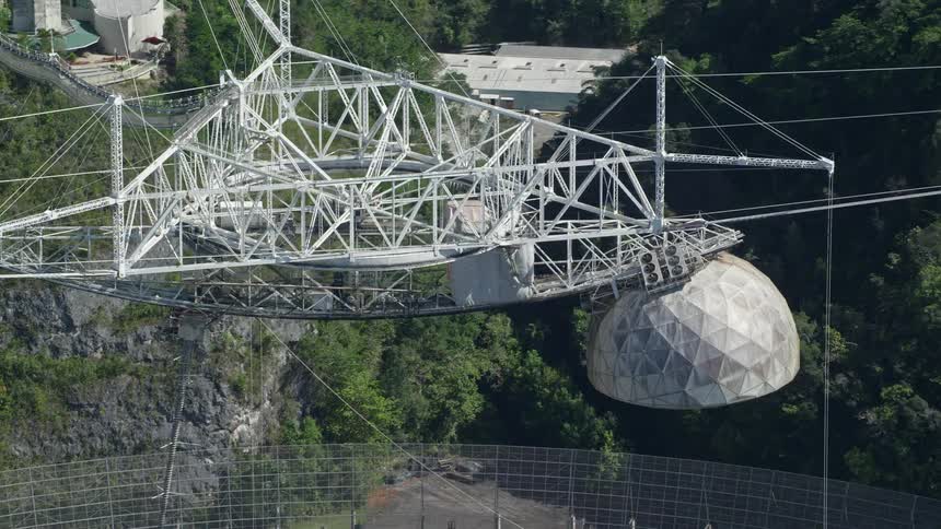 A giant radio telescope sits near buildings and a forest