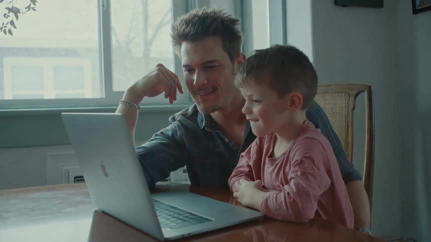 A man and a young boy sit at a table both laughing at a laptop