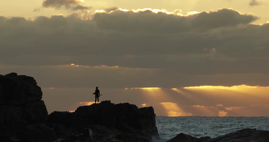 A surfer stands on rocks next to the ocean