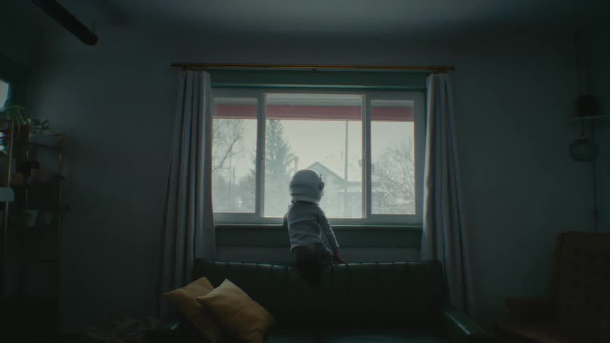 A young boy wearing an astronaut helmet looks out his living room window
