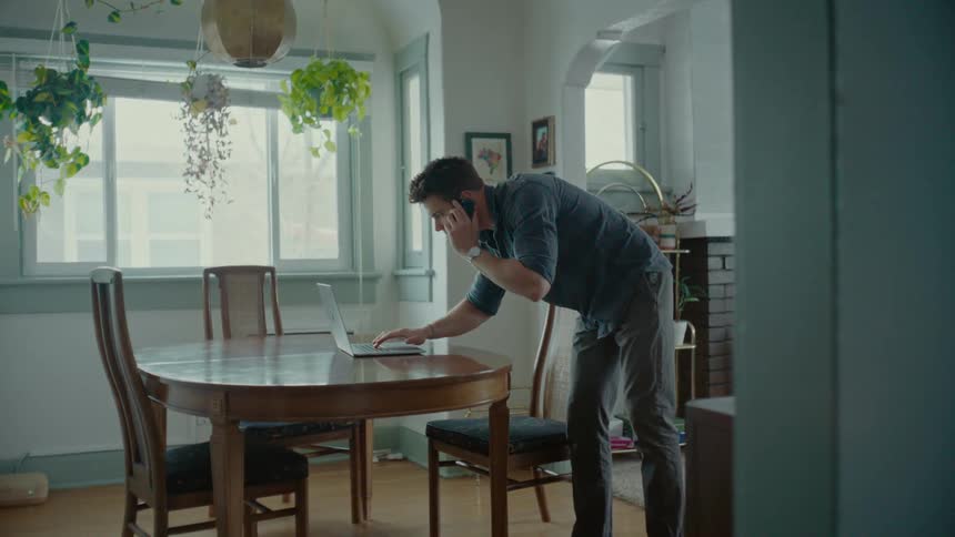 Father answering the phone and typing on the laptop in the living room with two children running around